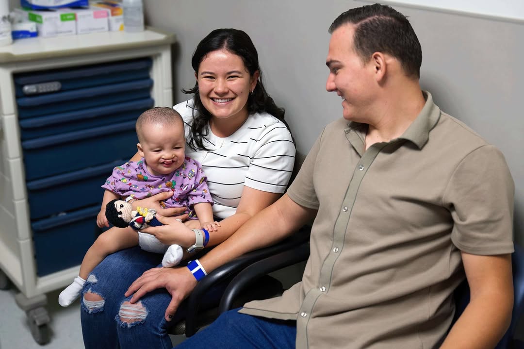 Smiling mother holding a happy baby with a toy, sitting next to a man in a medical office setting.