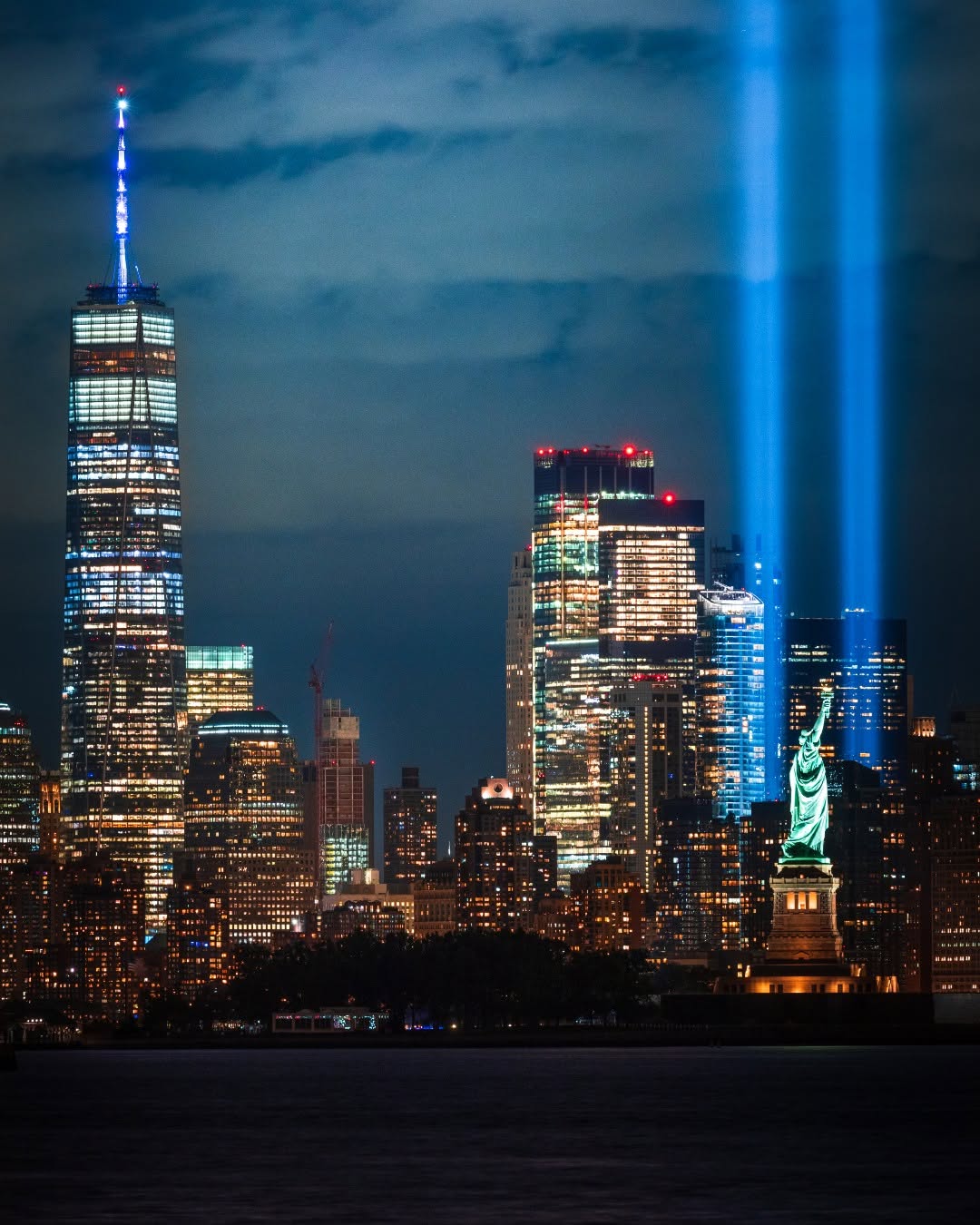 A nighttime view of the New York City skyline featuring the illuminated One World Trade Center and the Statue of Liberty. Twin beams of light also shine into the sky, symbolizing the Tribute in Light.