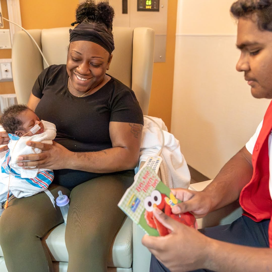Mother holding newborn in hospital room while a man reads a children's book to the baby.