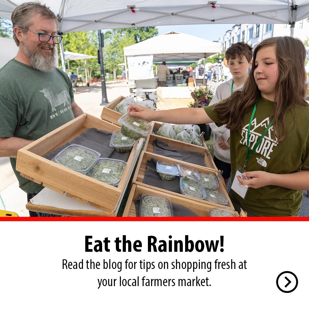 A young girl selects fresh greens from a vendor's wooden display at an outdoor farmers market under white tents.