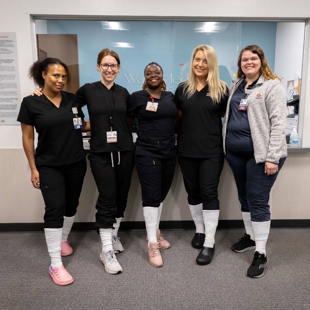Five healthcare workers in black and navy scrubs stand together smiling in a hospital hallway, wearing white protective shoe covers.