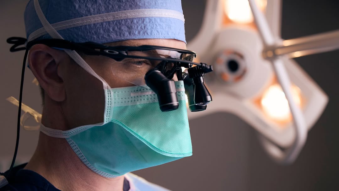 Close-up of a surgeon focusing while wearing a face mask and magnifying glasses under surgical lights.