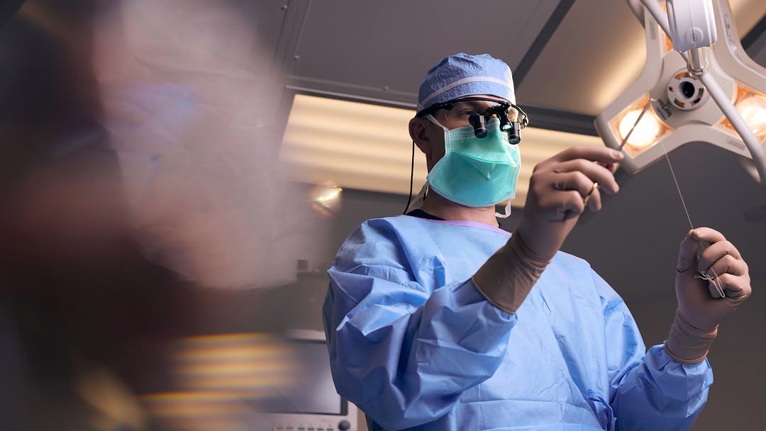 Surgeon in scrubs and mask working under surgical lights.