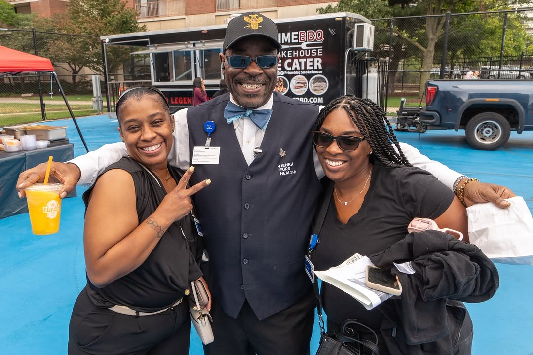 A group of three people stand smiling on a blue court. The person in the center wears a vest labeled "Strong Memorial Hospital" and a bowtie, with their arms around the others. Another person holds a 