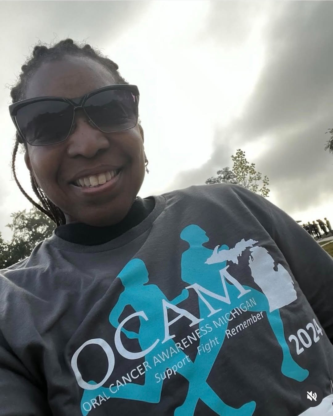 An individual is smiling and wearing sunglasses, dressed in a t‑shirt that reads "OCAM 2024 Oral Cancer Awareness Michigan Support. Fight. Remember." The background features a cloudy sky and trees.