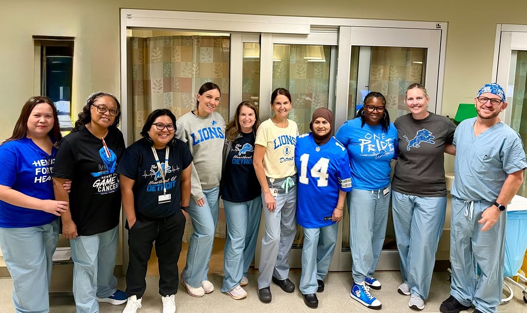 A group of healthcare professionals standing together in a hospital setting. They are wearing scrubs and various shirts, some featuring Detroit Lions logos. The backdrop includes a hospital curtain an