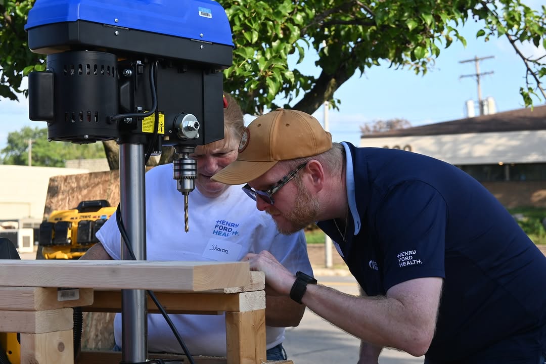 Two individuals are focused on a large power tool mounted on a wooden table outdoors. One person is wearing a cap and glasses. Both are engaged in adjusting or inspecting the equipment. In the backgro