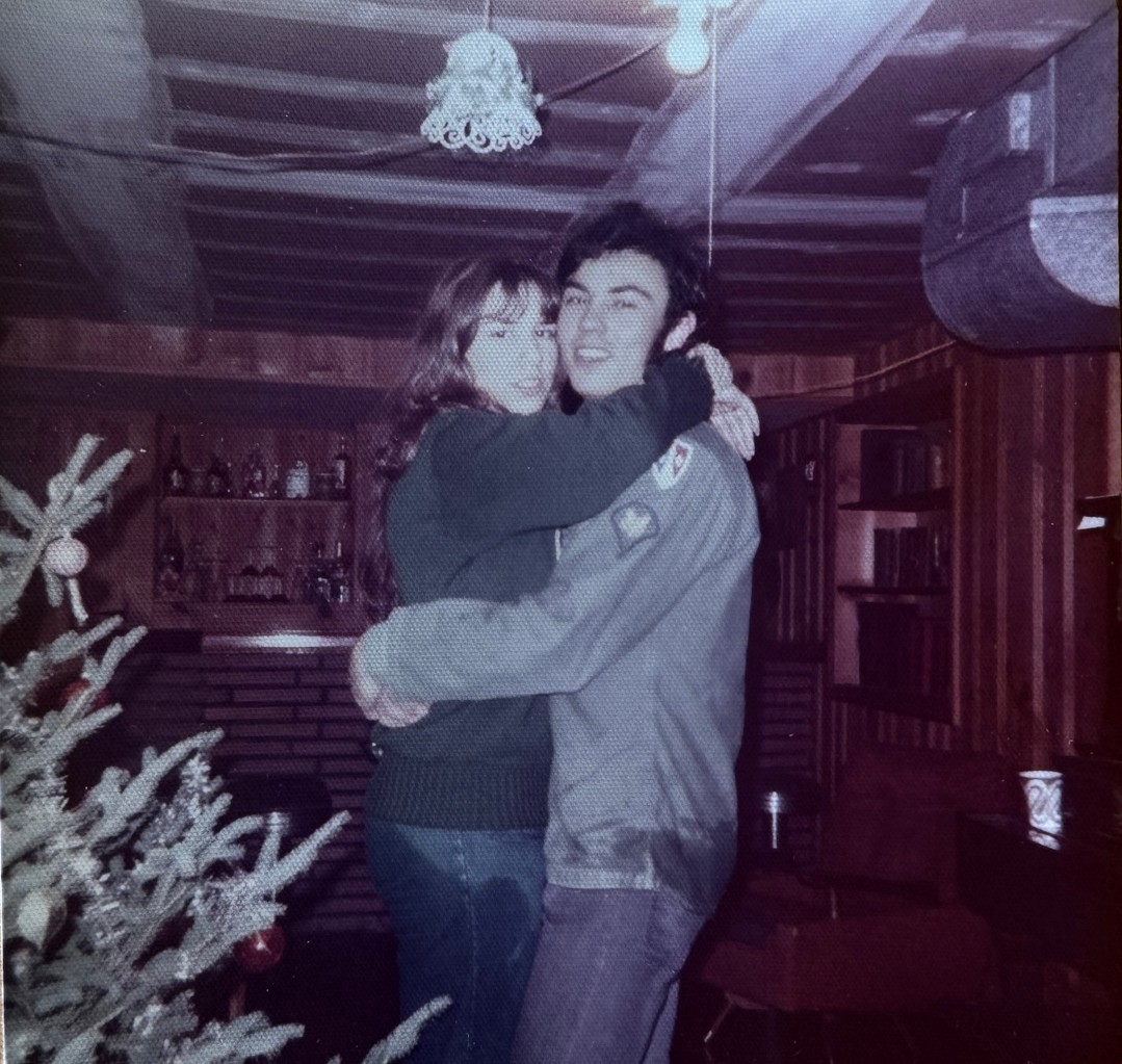 A person embraces another near a bar inside a room with wooden walls and a ceiling. A decorated Christmas tree is partially visible in the foreground.