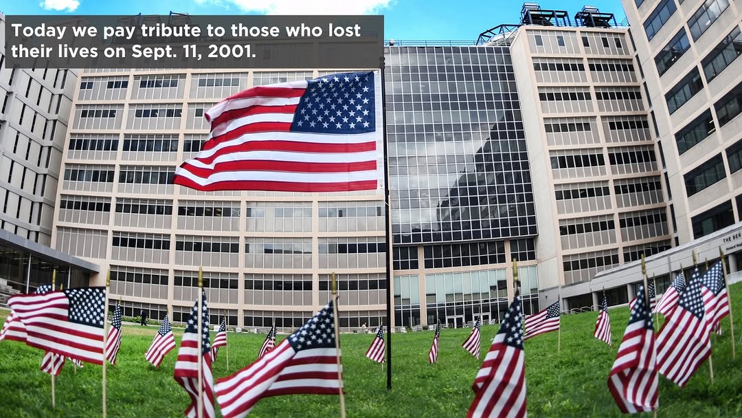 A display of American flags placed on a grassy area in front of a large modern building. A message reads: "Today we pay tribute to those who lost their lives on Sept. 11, 2001.