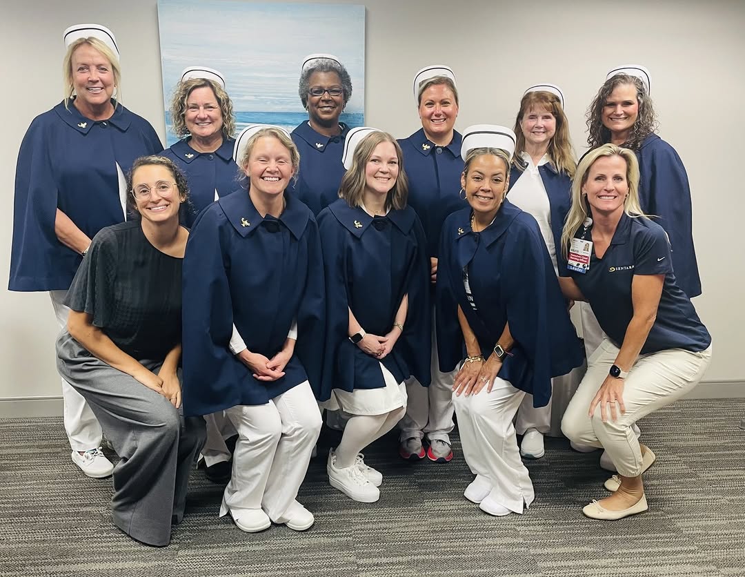 A group of people wearing navy blue capes and white nursing uniforms pose together in a room with beige carpeting and a light blue abstract painting on the wall. One person is wearing a badge. They ar