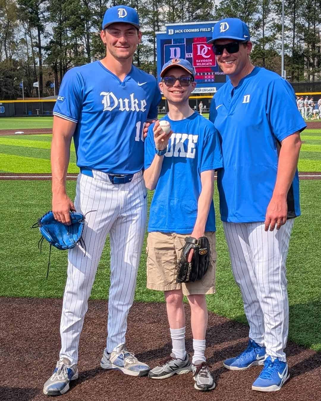 Three people are posing on a baseball field. Two are wearing blue "Duke" baseball uniforms, while the person in the middle (Alex Lucas) is wearing a blue "Duke" t-shirt and holding a baseball. Trees a