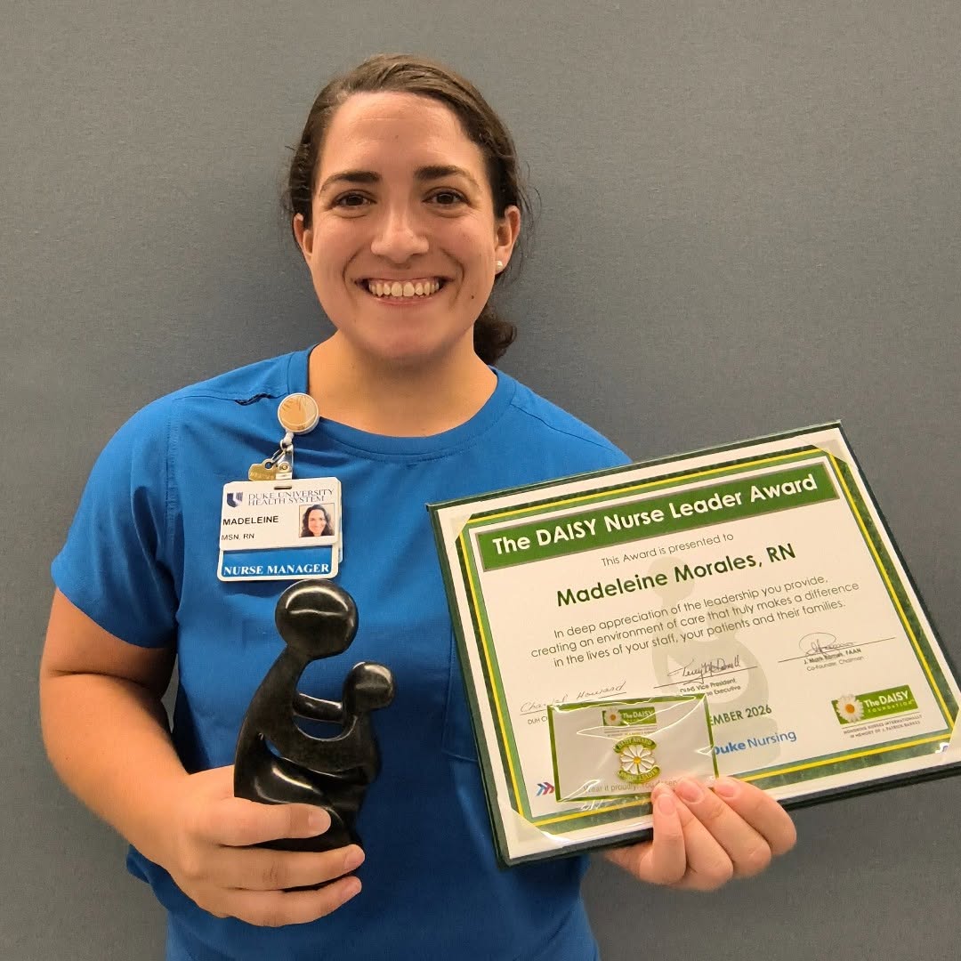A person in a blue uniform smiling at camera holding a certificate with "The DAISY Nurse Leader Award."