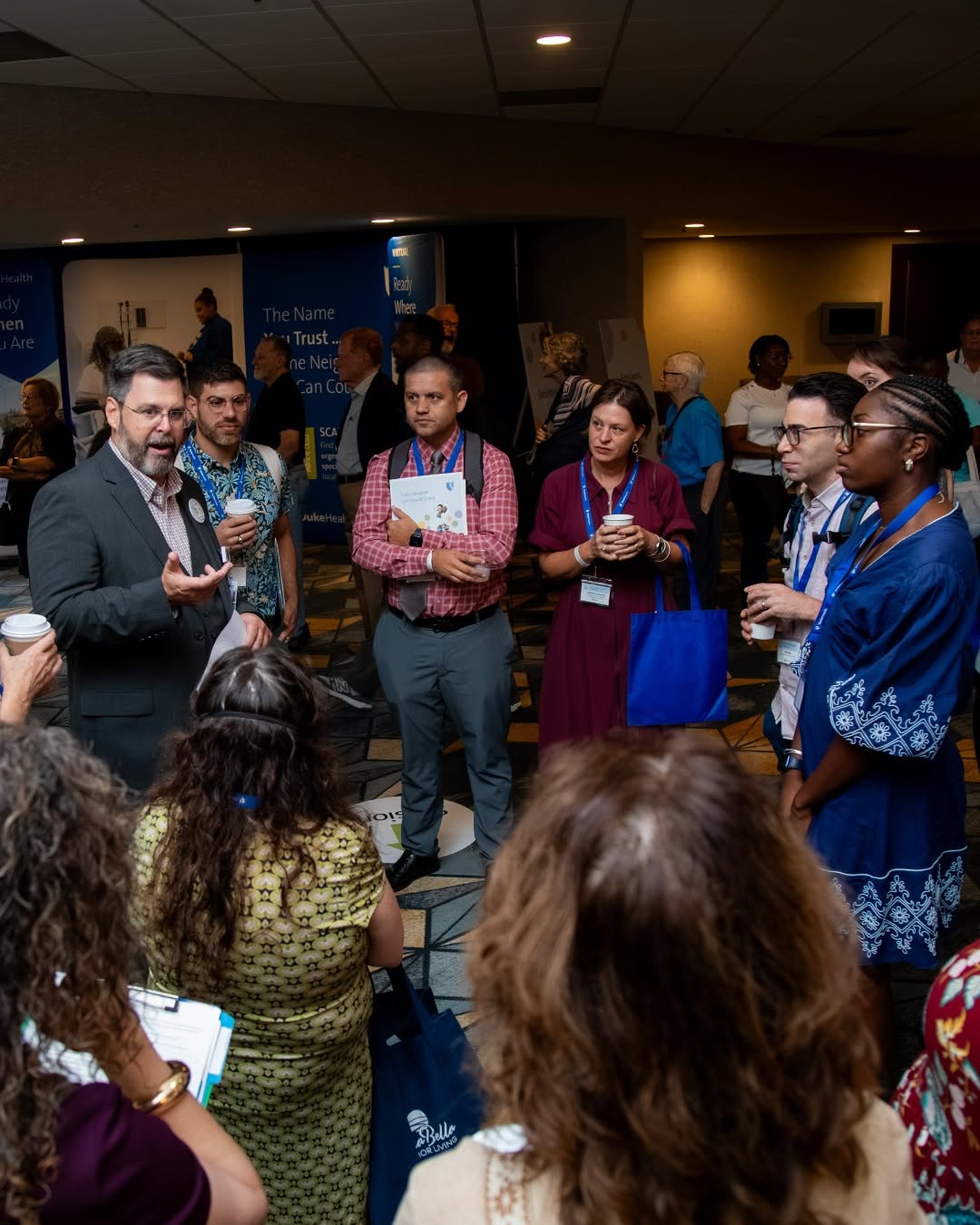 A group of individuals is gathered in a conference room, listening to a speaker holding a cup. The attendees are attentively engaged, standing in a semi-circle. Banners with legible text, including "W