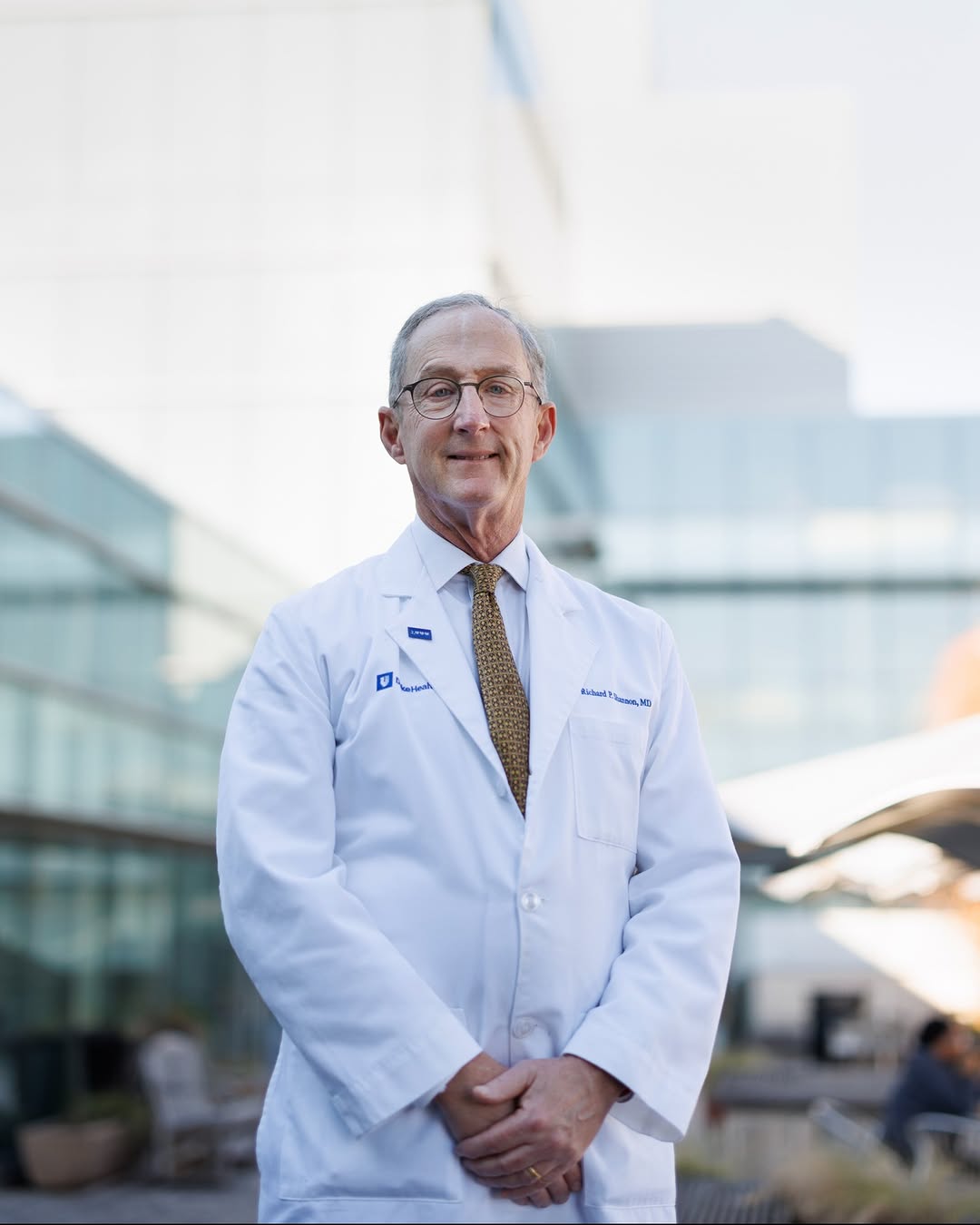 Richard P. Shannon, MD, in a white coat standing outside a modern glass medical facility building. The setting is well-lit with clear skies.