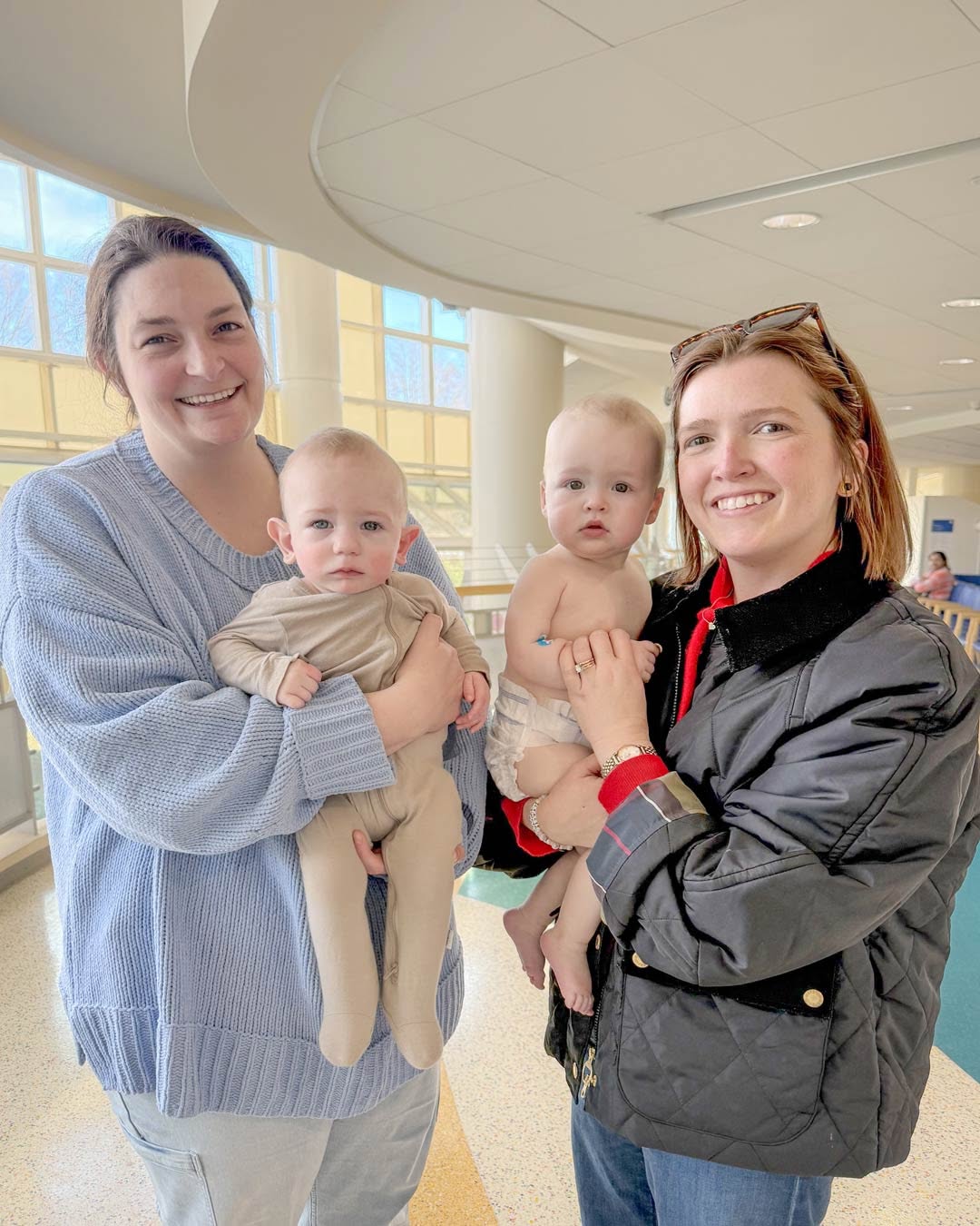 Two individuals are smiling while holding babies in their arms. They are indoors, with large windows in the background allowing natural light to fill the space at Duke Children's Hospital.