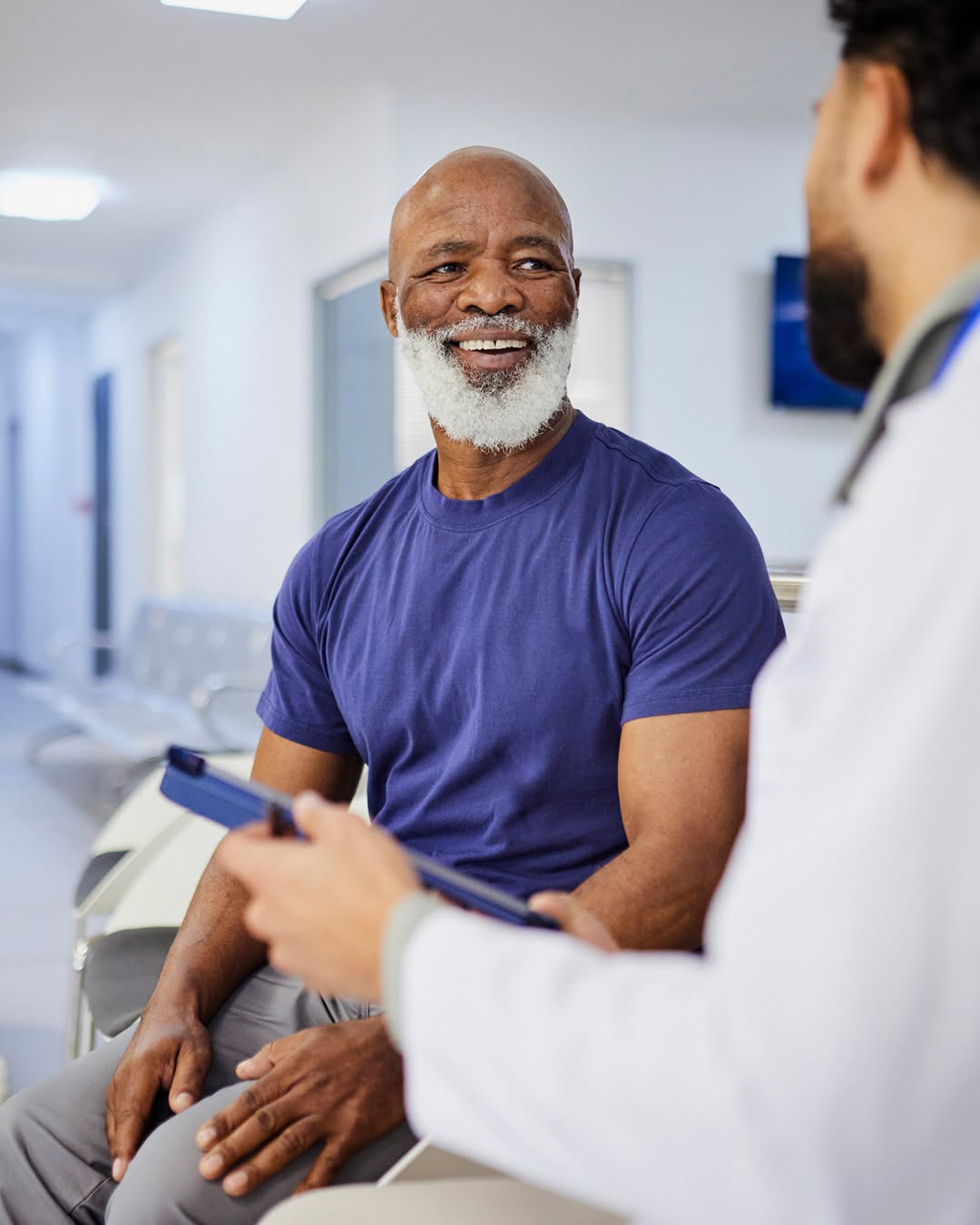 An individual with a white beard smiles while seated in a bright, modern clinic. Another person, in a white coat holding a clipboard, stands nearby, engaging in conversation. The setting suggests a me