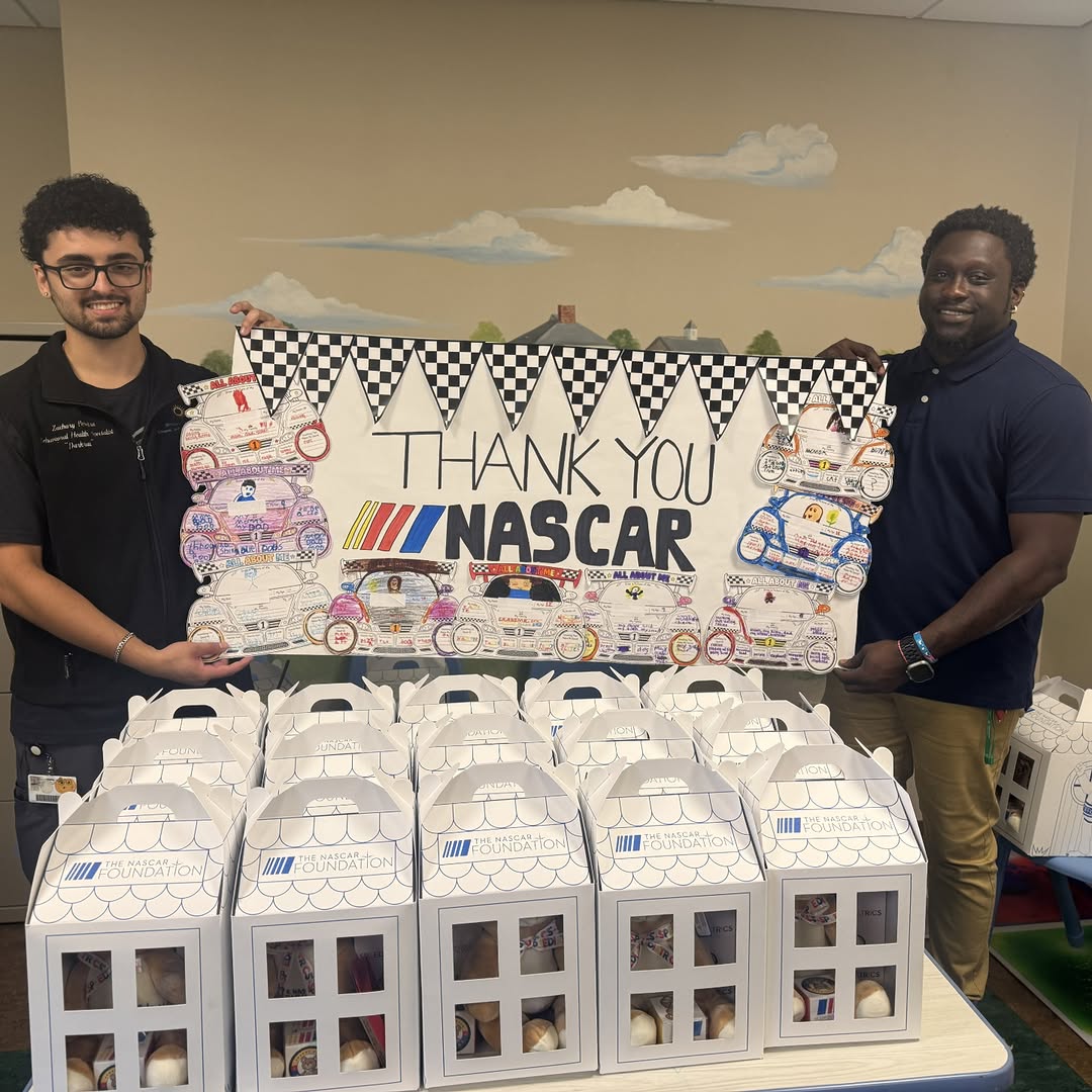 Two men holding a large "Thank You NASCAR" poster above rows of The NASCAR Foundation donation boxes.